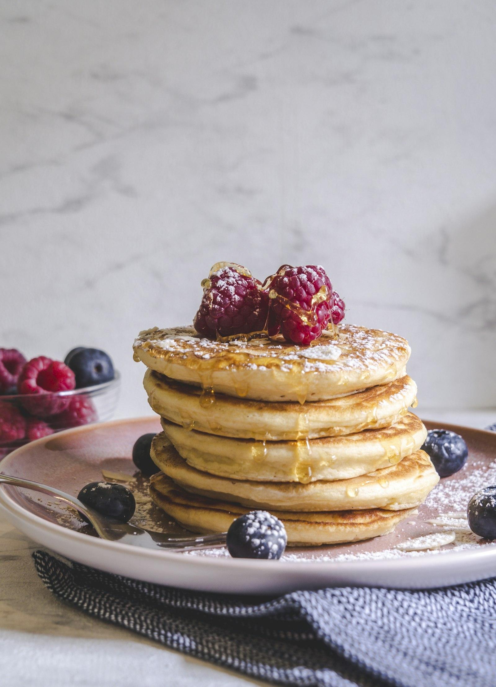 Tumpukan pancake fluffy dengan buah beri di atas piring putih, plating ala sarapan hotel
